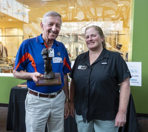 a man holding an alligator statue is staning next to a person in front of a huge cardboard display check in what looks like a public building like a library or museum