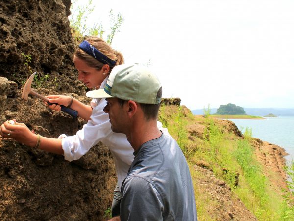 Two people stand next cliff face along a river bank. One person uses tools to dig while the other person watches.