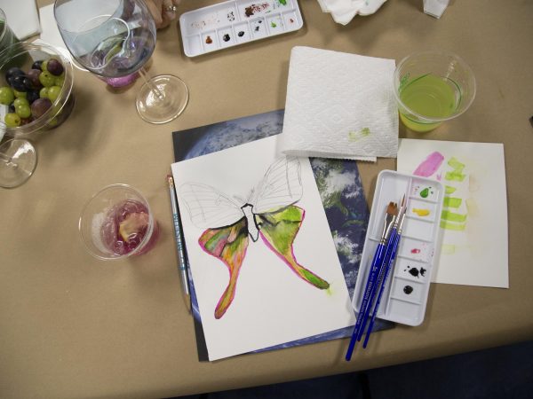 A half-finished painting of a moth. Around the painting are painting suppliers and a mocktail drink