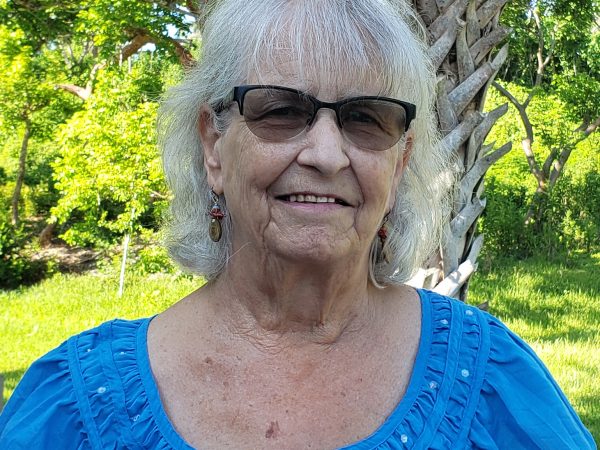 potrait of an older woman in a blue shirt, behind her are green trees and grass