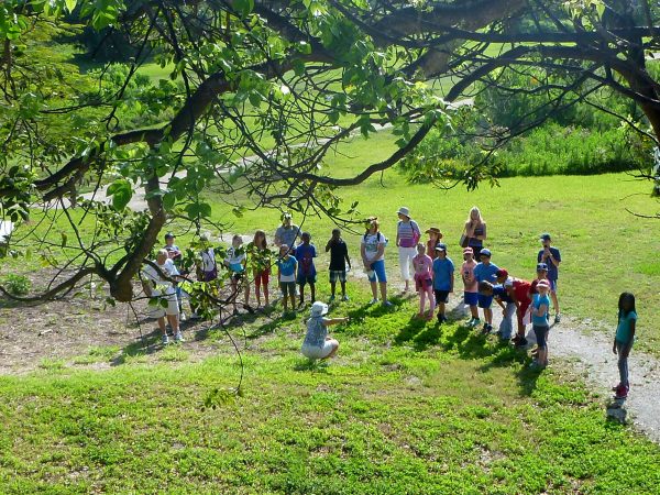 a person kneels at the bottom of a hill and gestures while they speak to a group of people gathered in a semicircle at the base of the hill